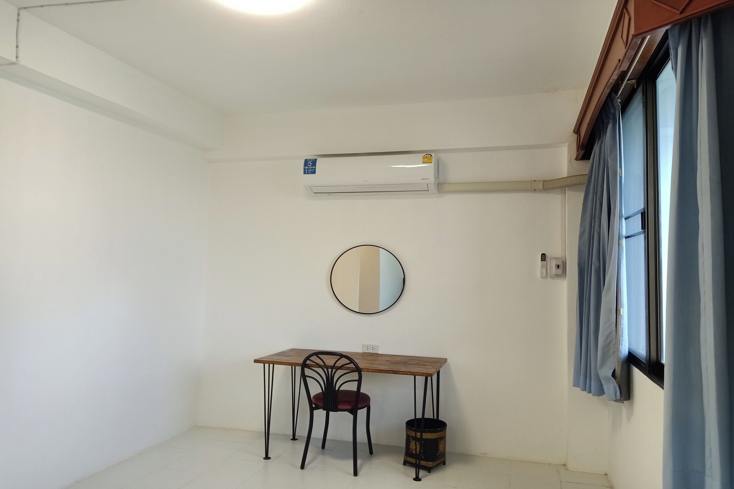 Et lille, rent værelse med et træskrivebord, en stol, et rundt spejl og en aircondition på væggen.