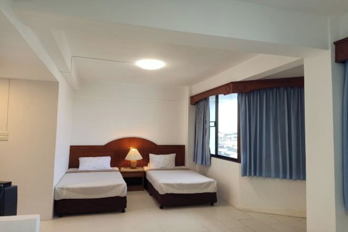 A hotel room with two single beds, a nightstand with a lamp, a window with blue curtains, and a view of the city.