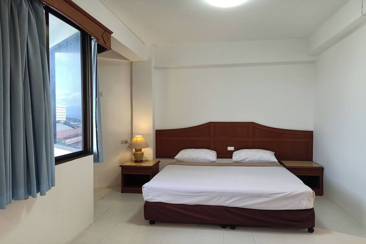 Una habitación de hotel con una cama grande, dos mesitas de noche, una lámpara y un ventanal con cortinas.