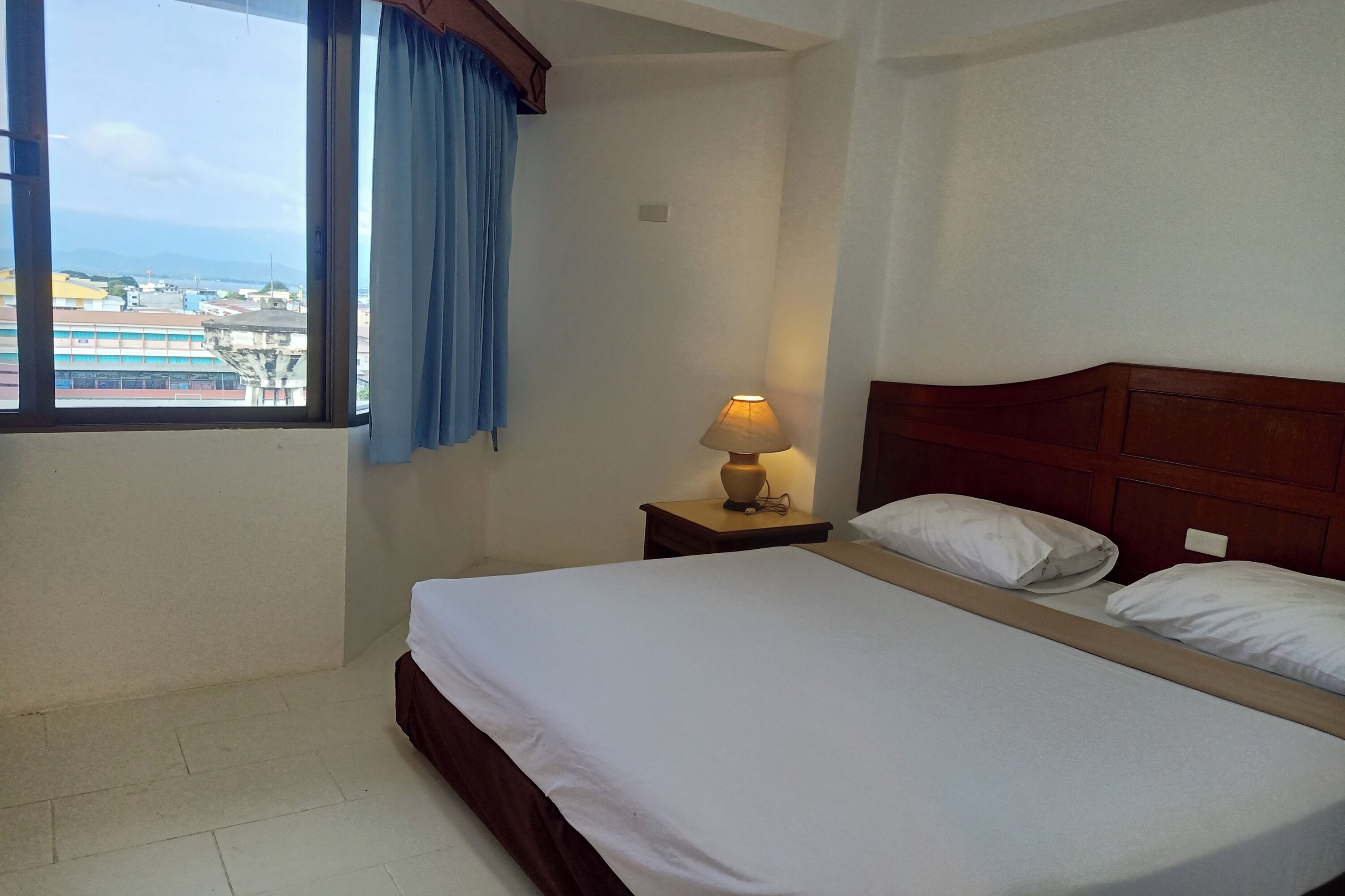 A hotel room with a bed, a nightstand with a lamp, a window with curtains, and a view of buildings outside.