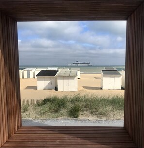 A view through a wooden-framed window overlooking a beach with white beach huts and a ship in the distance. - Comfortable seaside studio with terrace 200 m from the beach (Sangatte)