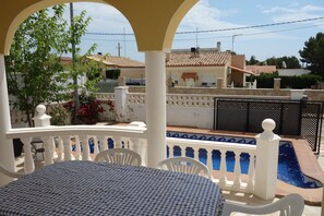 Un espace extérieur abrité comprenant une piscine, des chaises blanches et une table recouverte d’une nappe à carreaux.