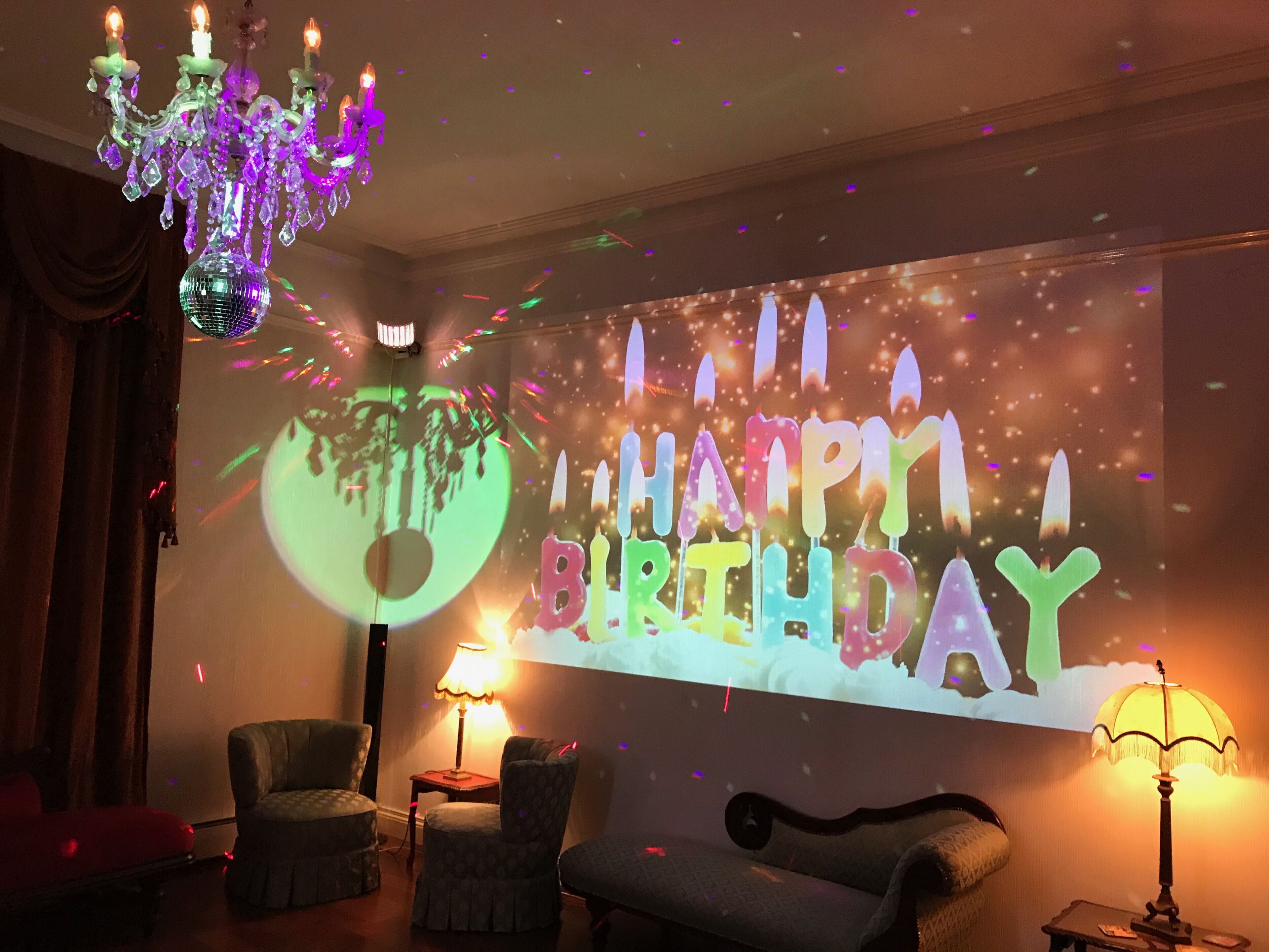 A room with a large screen displaying "HAPPY BIRTHDAY" and a disco ball. There are two armchairs and a lamp on a side table.