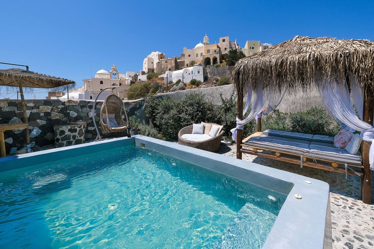 Una piscina con sillas de descanso y una sombrilla, rodeada de muros de piedra y vistas a edificios tradicionales.