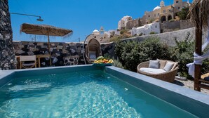 A swimming pool with a wicker chair and a table, surrounded by stone walls and a view of a white building with domes.