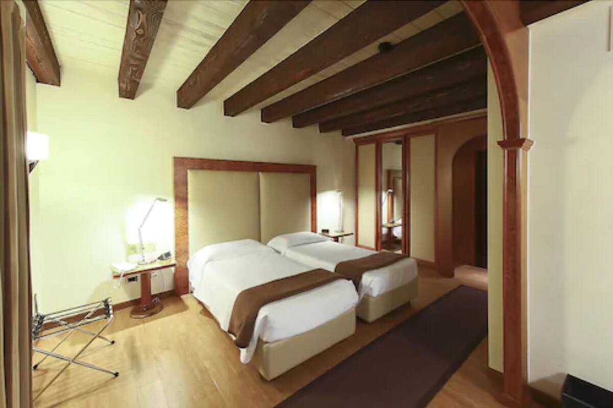 A hotel room with two beds, wooden beams on the ceiling, a bedside table with a lamp, and a wooden floor.