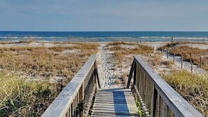 Una pasarela de madera que lleva a una playa con dunas y el océano al fondo.