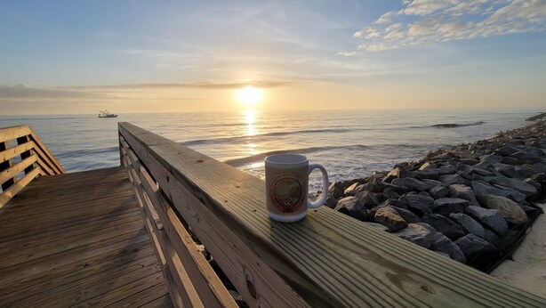 Une terrasse en bois avec une tasse posée sur une rambarde, donnant sur une côte rocheuse au coucher du soleil.