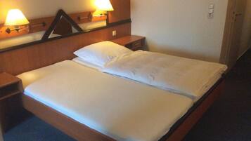 Een net opgemaakt bed met witte lakens in een hotelkamer. Er staan twee bedlampen op houten nachtkastjes, en een spiegel boven het bed.