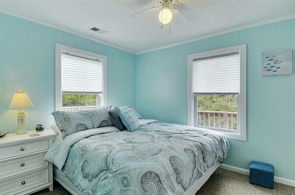 A bedroom with a bed, two windows, a nightstand, a lamp, and a small blue stool. - All Decked Out -OBX MP6 (Kill Devil Hills)