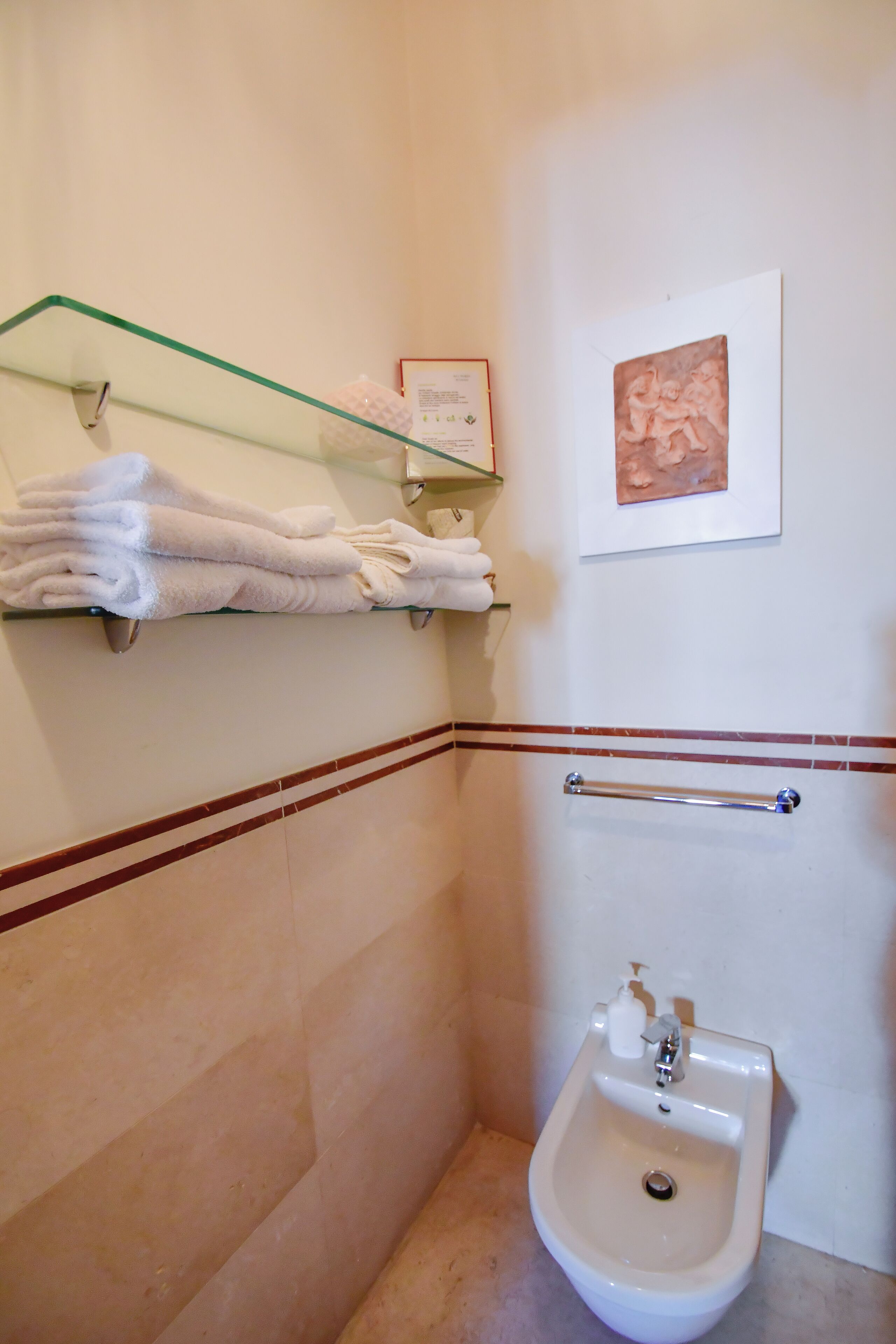 Un baño con repisas de vidrio que sostienen toallas dobladas, una obra de arte enmarcada en la pared, un bidé y una barandilla.