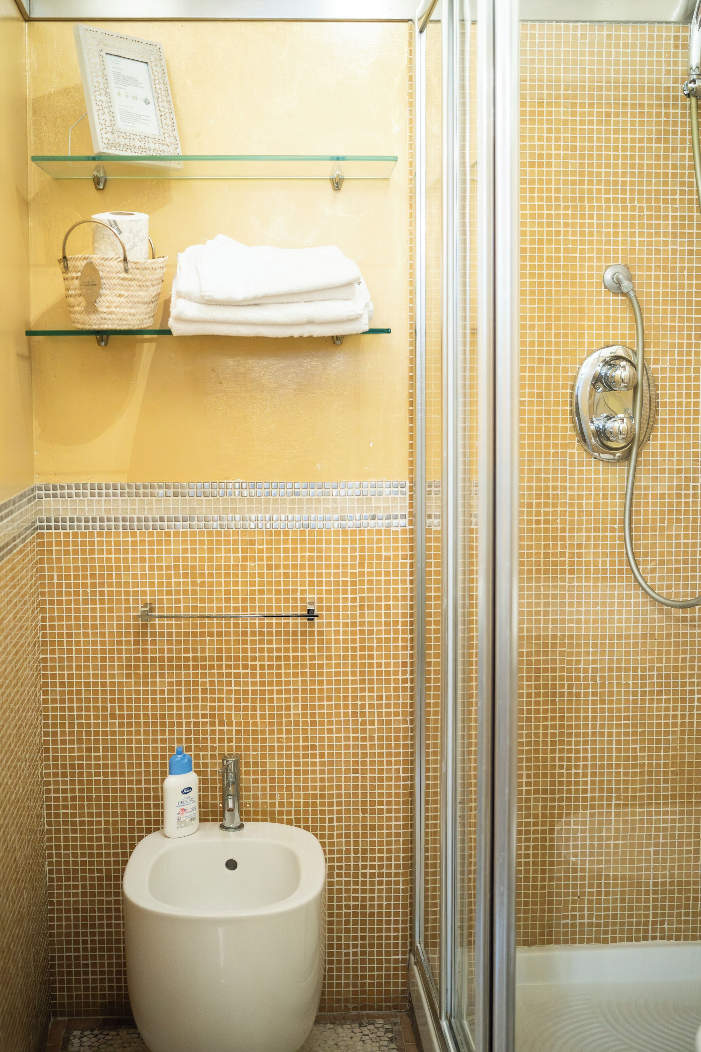 A bathroom with a glass shelf holding a towel and a basket, a bidet, and a shower area with a glass door.