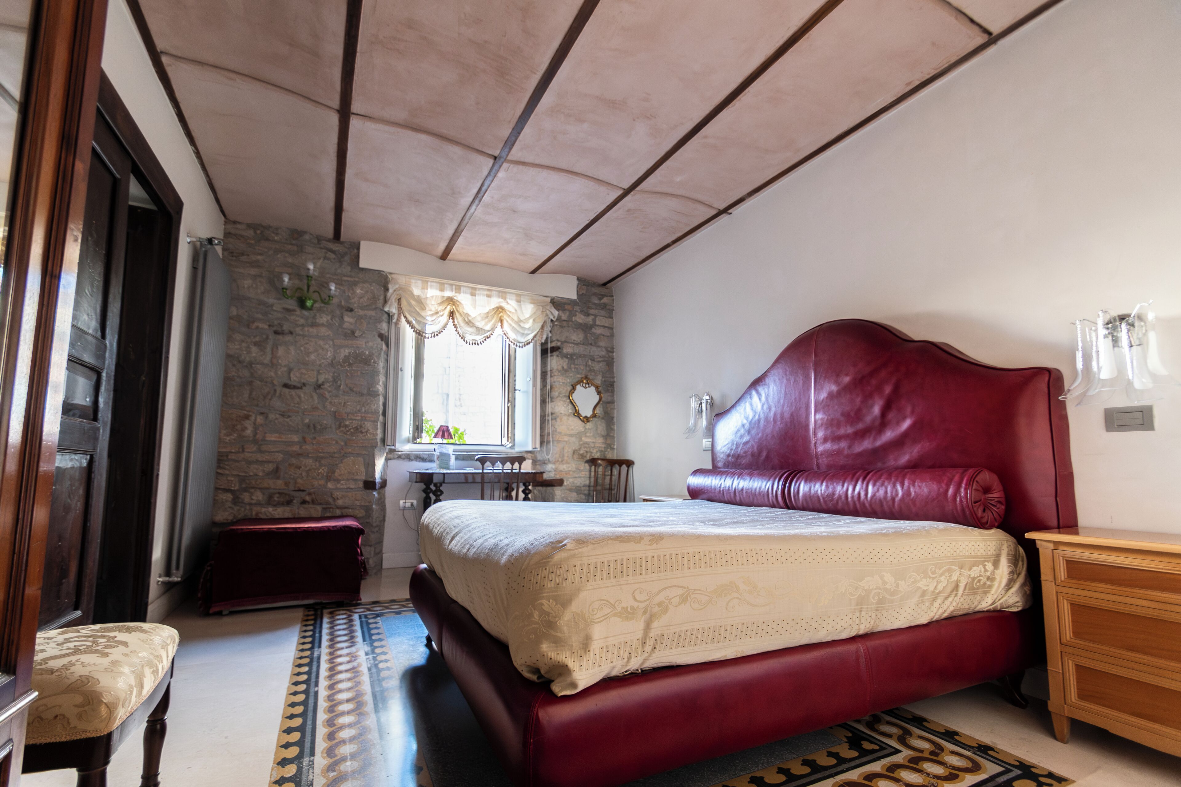 A bedroom with a large bed, a wooden nightstand, a bench, a stone wall, and a window with curtains.