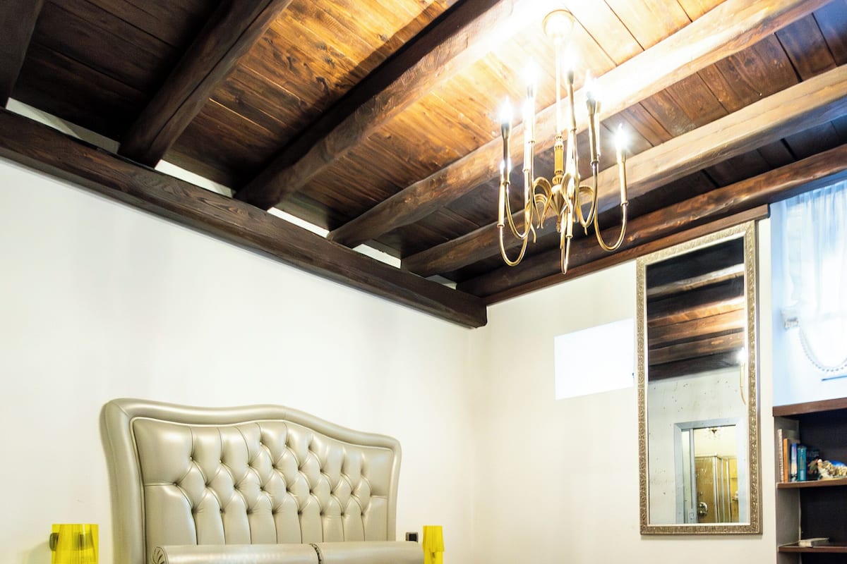 A bedroom with a tufted headboard bed, a patterned bedspread, a chandelier, a mirror, and a bookshelf.
