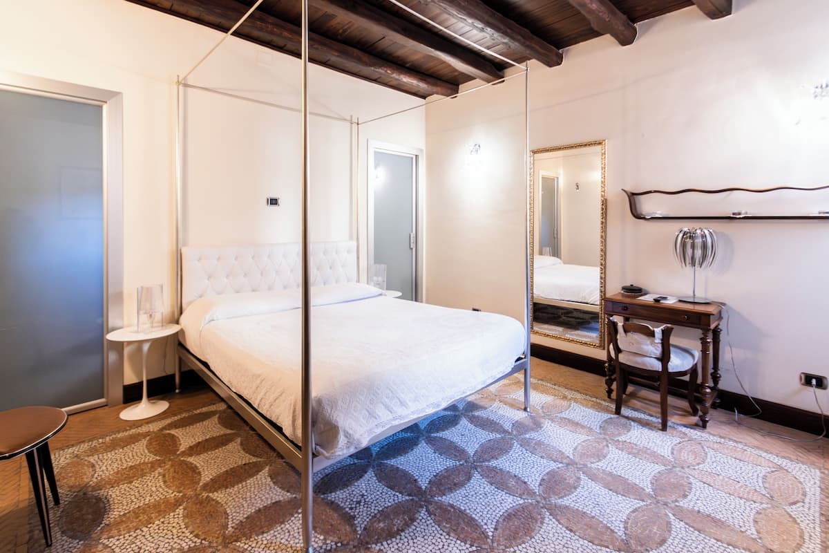 A bedroom with a four-poster bed, a bedside table, a chair, a mirror, and a small desk with a lamp.
