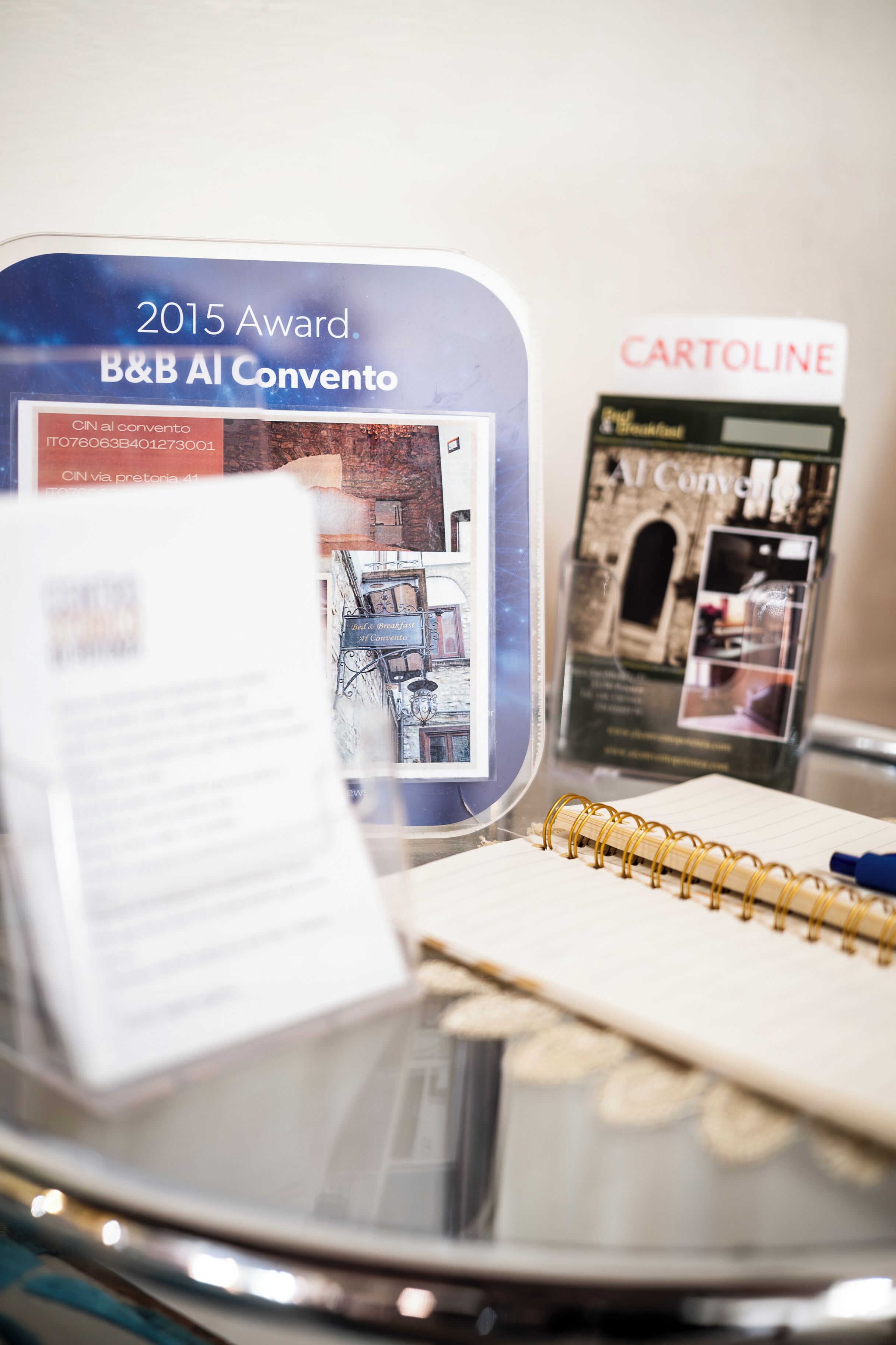 Una mesa de vidrio con un cuaderno, un folleto y un cartel para el B&B AI Convento, premio 2015.