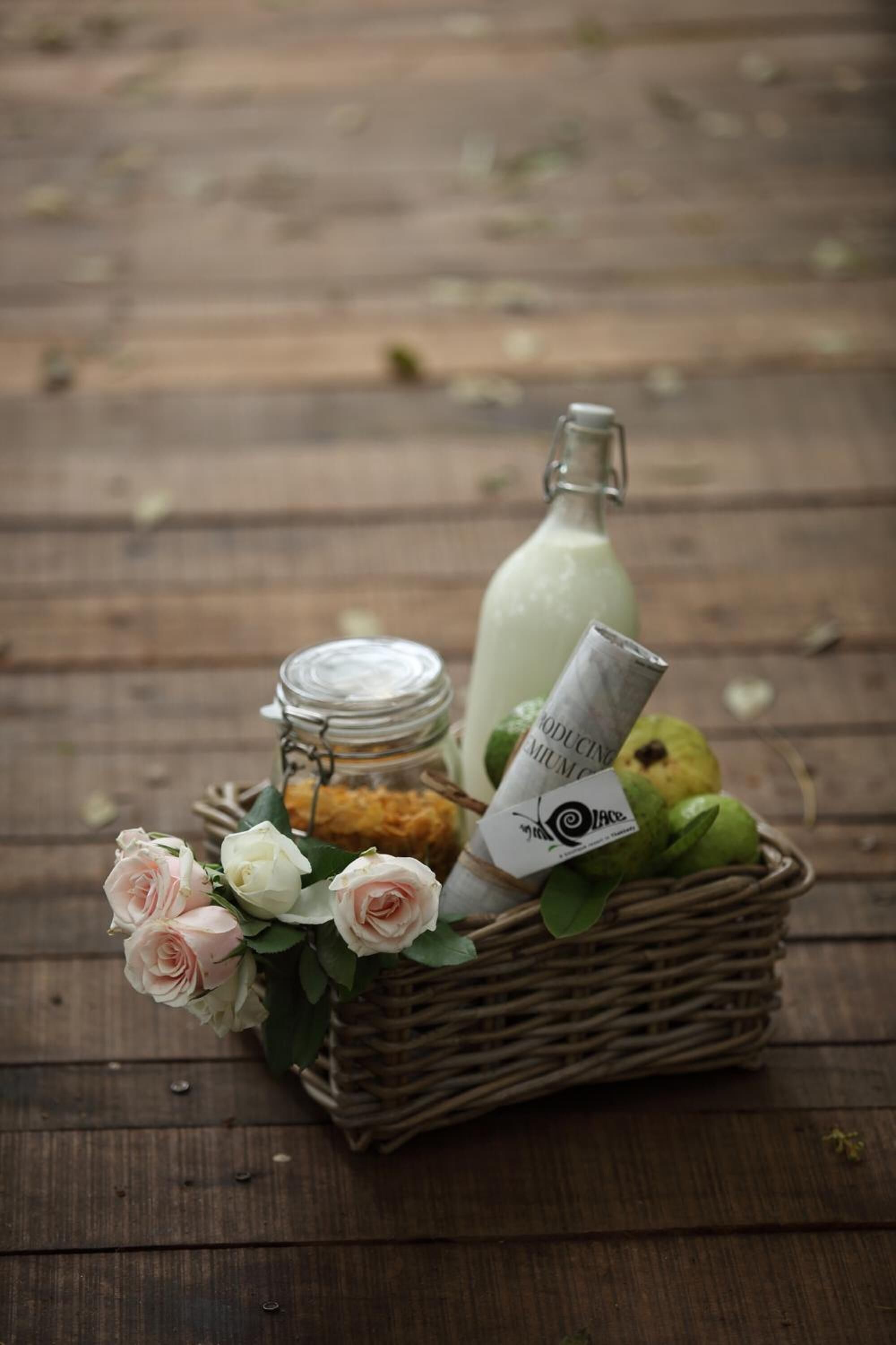 Un panier en osier contenant un pot de sucre jaune, une bouteille de liquide blanc, un objet enroulé avec du texte, et un bouquet de roses blanches et roses, le tout posé sur une surface en bois.