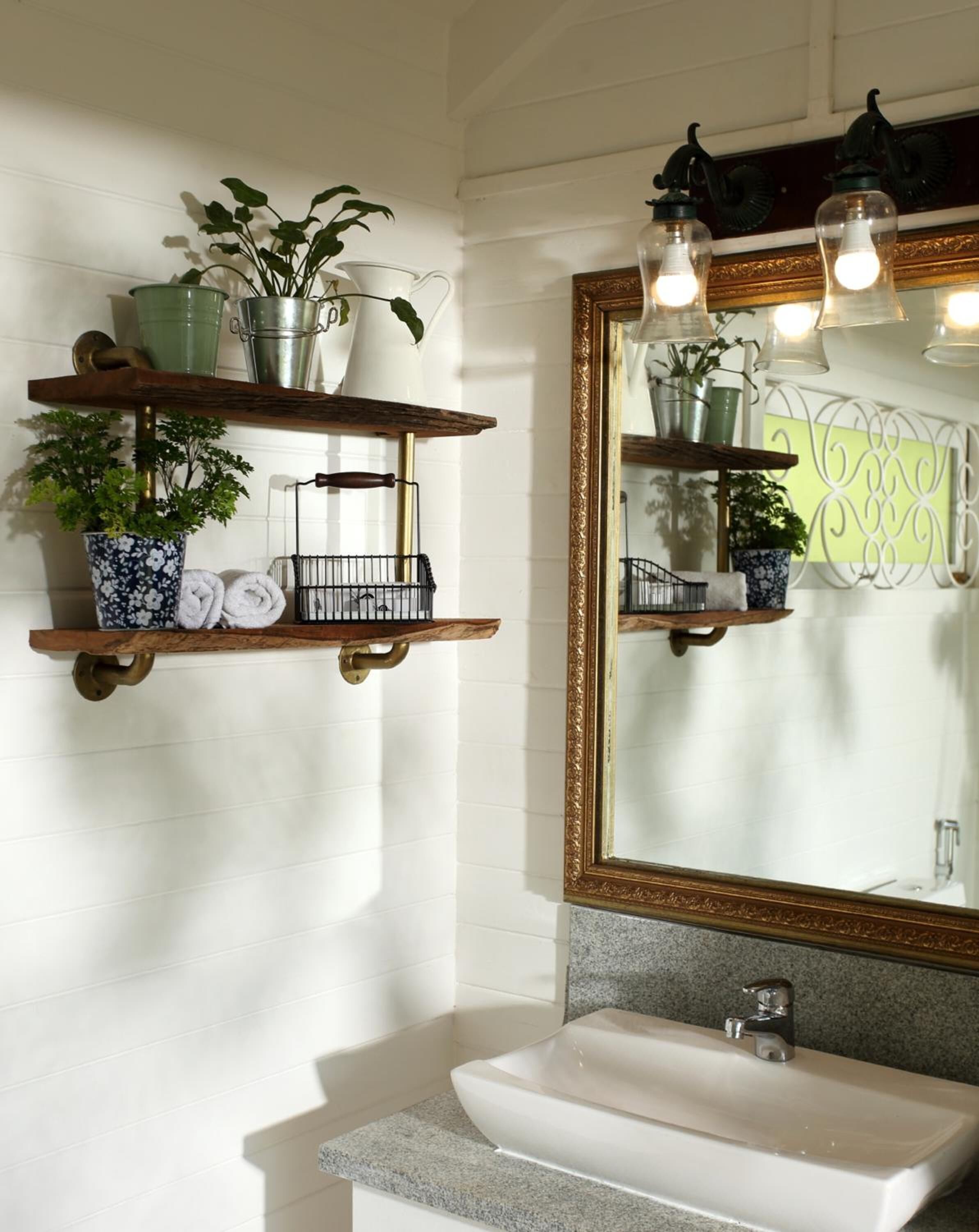 Un baño con azulejos blancos tipo metro, un lavamanos blanco y un espejo con marco dorado. Se observan estantes de madera con plantas y toallas, y una lámpara sobre el espejo.