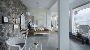 A spacious bedroom with a stone wall, a large bed, and a balcony with wicker furniture. - Hills & Hues (Peermade)