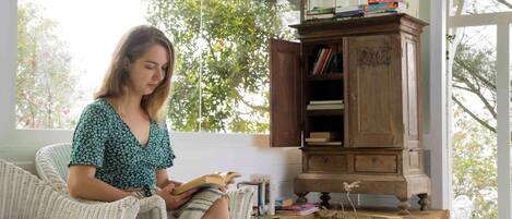 A person sitting on a wicker chair reading a book in a room with a wooden cabinet and a small table with books.