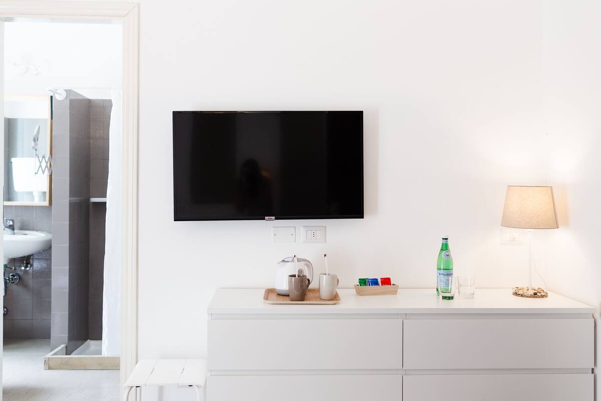 Une salle de bain moderne avec un meuble-lavabo blanc, un téléviseur à écran plat fixé au-dessus, un verre d’eau, une bouteille de Sprite et une lampe sur le meuble-lavabo.