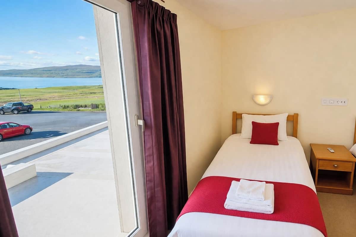 A hotel room with two beds, a window with curtains, a view of a road and car, and a small bedside table.