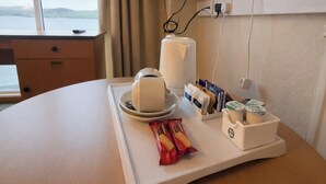 Habitación de hotel con una mesa de madera, una bandeja con una taza, un control remoto, un televisor montado en la pared y una ventana con vista al mar.