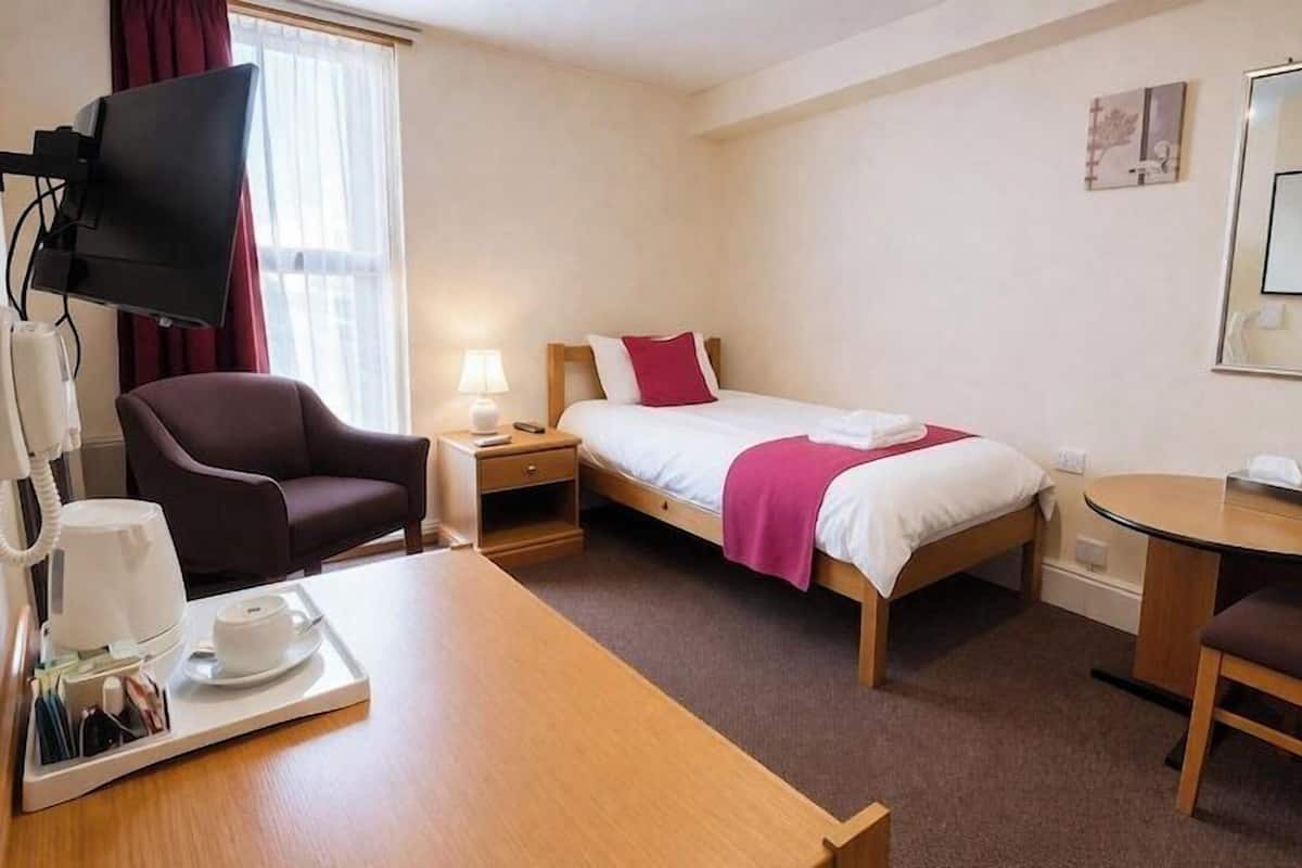 A hotel room with a bed, a desk, a chair, a TV, a bedside table, and a window.