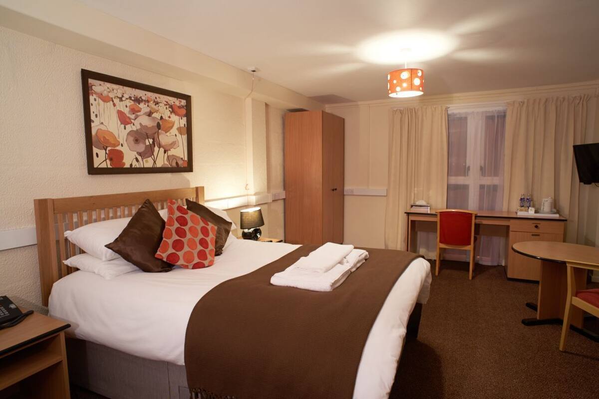 A hotel room with a bed, a desk, a chair, a lamp, and a painting on the wall.