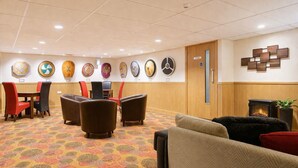 Un hall de hotel con alfombra estampada, un área de descanso con sofás y sillones, una chimenea y relojes en la pared.