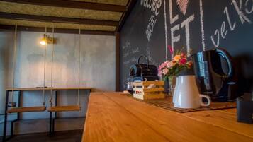 Ein Holztisch mit einer Kaffeemaschine, einer Tasse und einem Brotkorb. Eine Schieferwand mit den Aufschriften 'WELCOME' und 'VELK'.