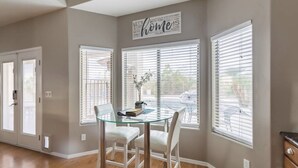 Uma área de jantar com mesa e cadeiras de vidro, uma placa decorativa acima das janelas e vista para a rua.