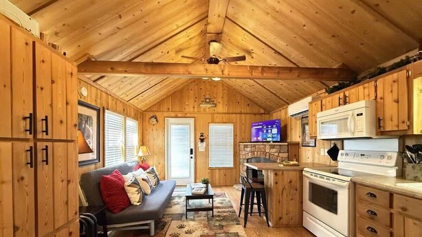 A cozy cabin interior with a wooden ceiling, a kitchen area with a stove and microwave, a dining table, and a sofa with cushions. - Mountain Views | Fire Pit | little bear cabin (Island Park)