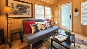 A cozy cabin interior with a wooden ceiling, a sofa with patterned cushions, a small table with books, and a framed picture on the wall. - Mountain Views | Fire Pit | little bear cabin (Island Park)