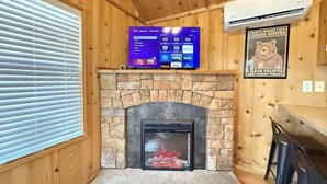 A room with a stone fireplace, a mounted TV displaying various channels, and a bear-themed poster on the wall. - Mountain Views | Fire Pit | little bear cabin (Island Park)