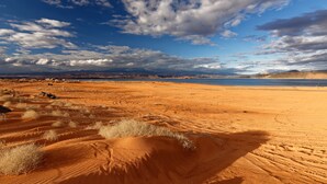 Wüstenlandschaft mit Sanddünen und einem fernen Gewässer unter einem teils bewölkten Himmel.