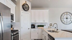 Eine moderne Küche mit weißen Schränken, einem Edelstahl-Kühlschrank, einem Gasherd, einer Kaffeemaschine und einer Wanduhr.