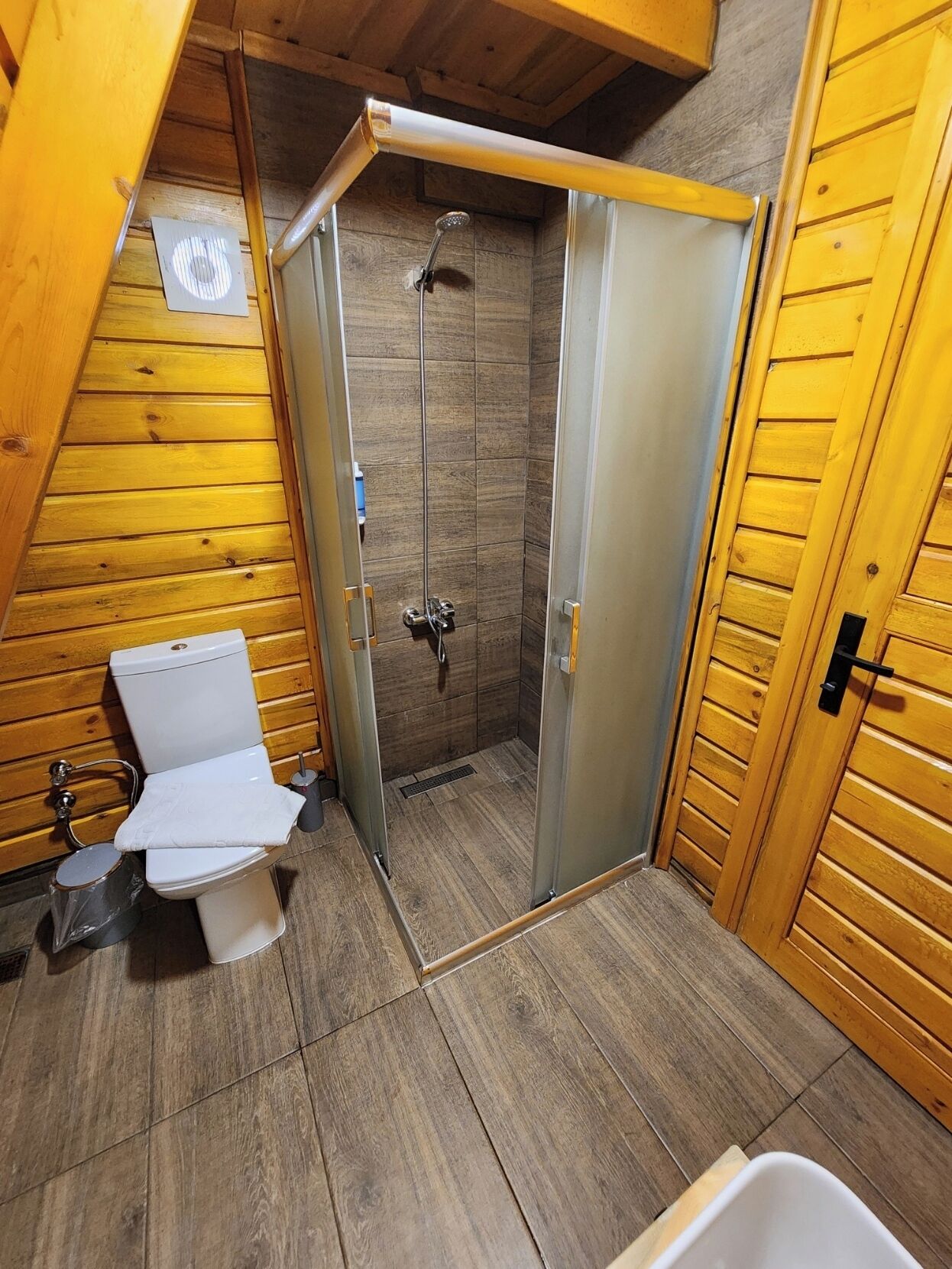 A small, wooden-paneled bathroom with a shower, toilet, and sink.