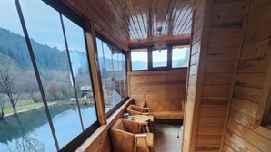 Une cabane en bois dotée de grandes fenêtres donnant sur un lac et des collines. À l’intérieur, on trouve un banc en bois et un canapé.