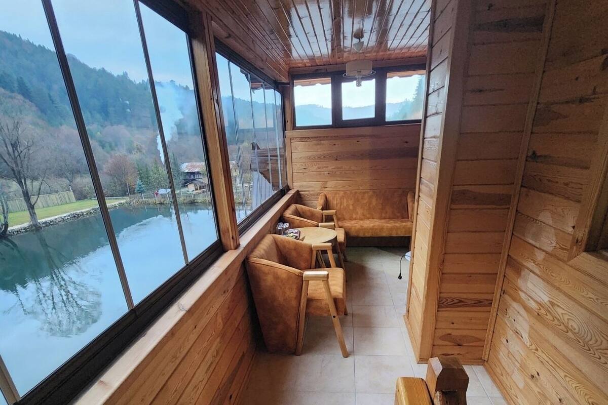 Uma cabana de madeira com grandes janelas voltadas para um lago e colinas. No interior, um banco de madeira e um sofá.
