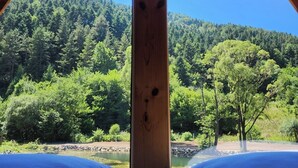 Une pièce avec un plafond en bois et un lit recouvert de draps blancs. Une grande fenêtre offre une vue sur une colline boisée et un plan d’eau.