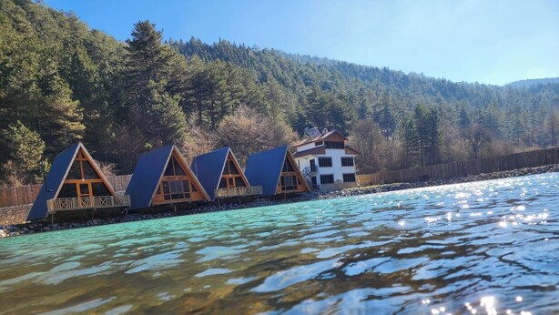 Une rangée de chalets en bois en forme de « A », avec un bâtiment blanc au centre, situés au bord d’un lac aux eaux claires, et une colline boisée en arrière-plan.