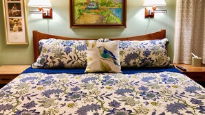 Una cama bien hecha con ropa de cama floral, dos lámparas de pared, un cuadro de paisaje enmarcado y una fotografía enmarcada en la pared.