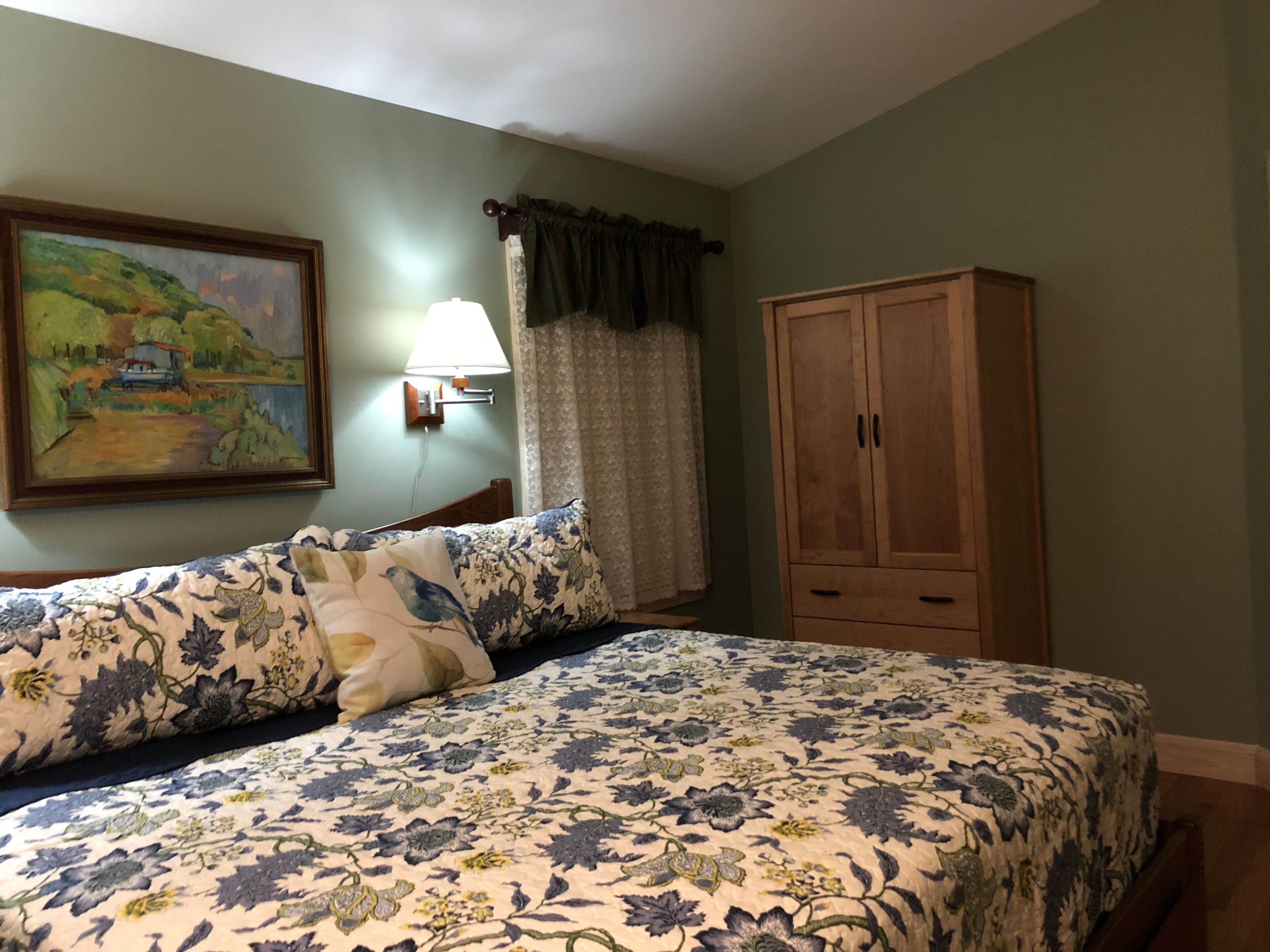 A bedroom with a floral bedspread, a wooden wardrobe, a painting on the wall, and a nightstand with a lamp.