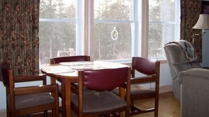 Un comedor con mesa y sillas de madera, ventanas amplias con cortinas, y una lámpara sobre una mesita.
