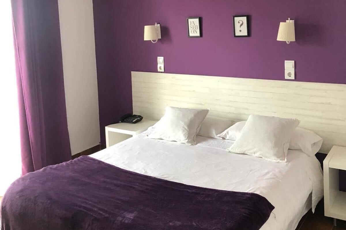 Un dormitorio con pared morada, cama blanca y dos mesitas de noche con lámparas.