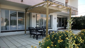 A covered outdoor seating area with chairs and tables, large windows, and a potted plant with yellow flowers.
