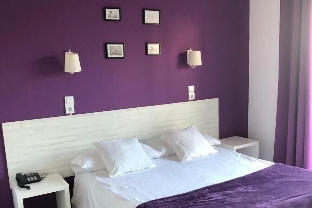 Una habitación de hotel con cama, mesitas de noche blancas, teléfono y lámparas fijadas a la pared.