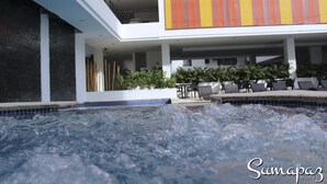 Et område med jacuzzi og en pool i baggrunden, samt en overdækket udendørs siddeplads og en bygning med en stribet facade.