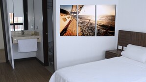 Et hotelværelse med en seng, et natbord, trægulv, håndvask på væggen og et håndklæde hængende på døren.